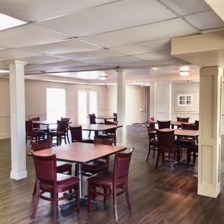An empty dining area with multiple tables and chairs, featuring wood flooring and ample natural light.