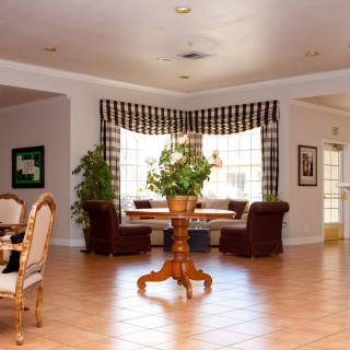 A well-lit living room with wooden floors, featuring a central table surrounded by chairs and green plants near the windows.