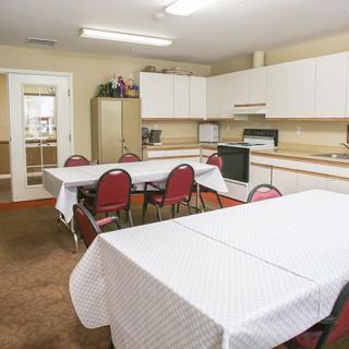 A kitchen-meeting room setup with tables covered in white tablecloths, red chairs, and kitchen appliances.