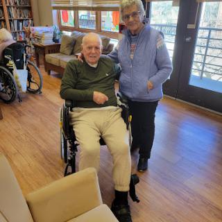 A caregiver assists an elderly man in a wheelchair in a welcoming community space.