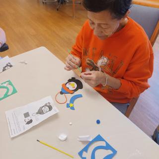 An elderly individual engages in a crafting activity, working on colorful materials at a table.