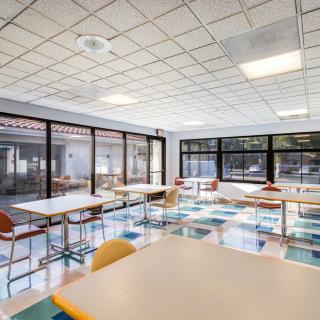 A bright classroom with tables and chairs arranged under large windows.