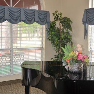 A grand piano next to a window with blinds and curtains, decorated with a vase of flowers.