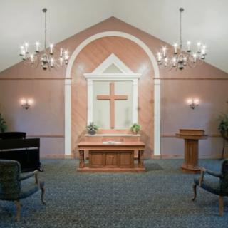 A serene chapel with a wooden altar and cross, flanked by chandeliers and seating.