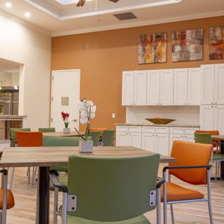 A modern dining area with colorful chairs and tables, featuring a kitchenette and decorative artwork on the wall.