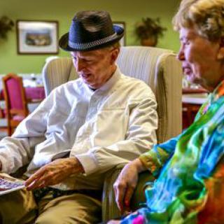 Two elderly individuals sit together, engaged in reading and conversation in a cozy indoor setting.