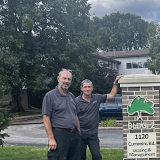 Two men stand beside a business sign in a lush, green environment.
