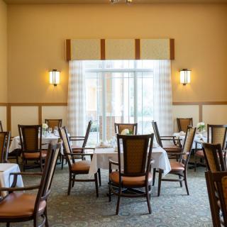A well-lit dining area with arranged tables and chairs, featuring elegant lighting and large windows with curtains.