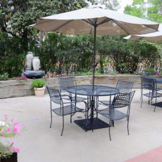 A serene outdoor patio with metal chairs, a table, and a large umbrella surrounded by greenery.