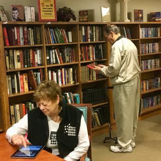 Two people in a library, one browsing books and the other using a tablet.