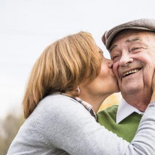 An elderly woman affectionately kisses an elderly man on the cheek, both smiling and enjoying a moment together outdoors.