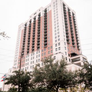 A view of a tall, modern skyscraper with a mix of white and pinkish hues, surrounded by trees in an urban setting.