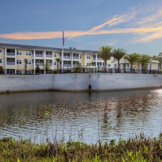 A scenic view of an apartment building by a lake with trees and a clear sky.