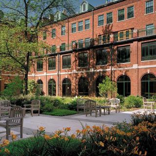 A brick building with arched windows overlooks a garden with benches and trees.