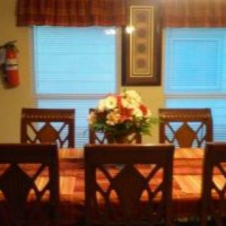 A cozy dining room with a flower centerpiece on the table, surrounded by chairs and illuminated by natural light from the windows.