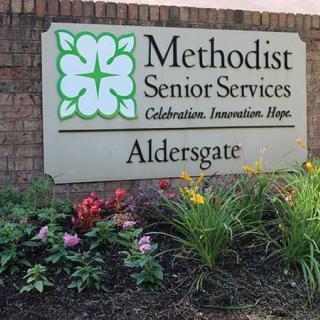 A sign for Methodist Senior Services at Aldersgate, surrounded by colorful flowers and a brick wall.