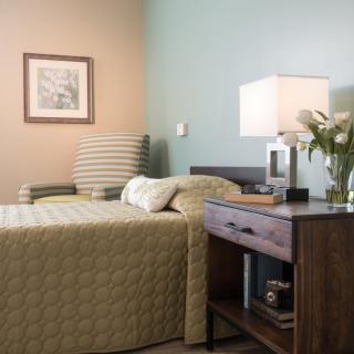 A cozy bedroom with a neatly made bed, a nightstand with a lamp and flowers, and a striped armchair.