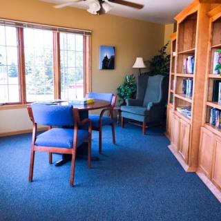 A cozy living room with wooden furniture, including a bookshelf and a table with chairs, illuminated by natural light from a large window.