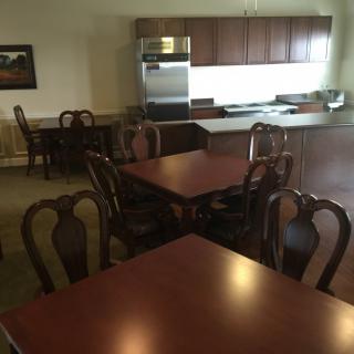 A dining area with wooden tables and chairs, adjacent to a kitchen with cabinets and appliances.