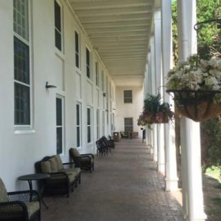 A long outdoor porch with columns and chairs lined up alongside hanging plants.