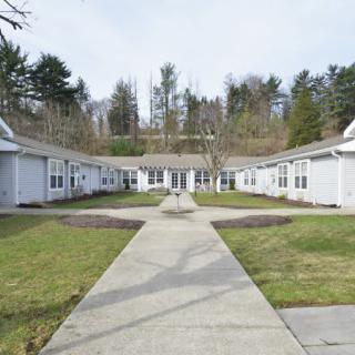 A courtyard surrounded by buildings with a central concrete pathway and grassy areas, set against a backdrop of trees and a clear sky.