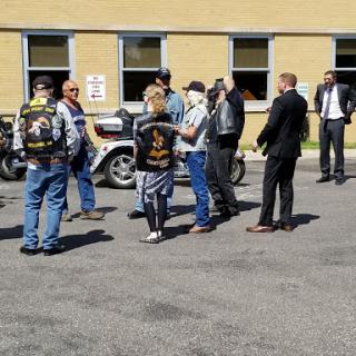 A diverse group of individuals, including bikers and formally dressed people, gathered outside near motorcycles.