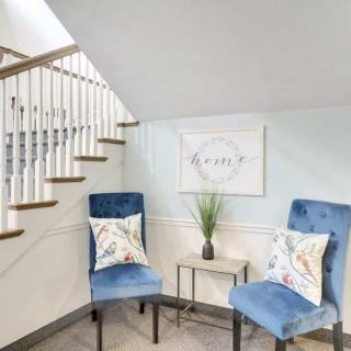 A cozy corner features two blue chairs with decorative pillows, a small side table with a plant, and wall art under a staircase.