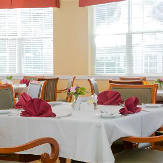 A neatly set dining room with tables featuring red napkins and wooden chairs, bathed in natural light from large windows.