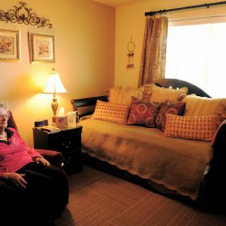 A cozy living room with an elderly woman sitting in a chair, surrounded by decorative cushions and a warm ambiance.