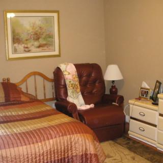 A cozy bedroom with a bed, armchair, and nightstand featuring a lamp and framed art on the wall.