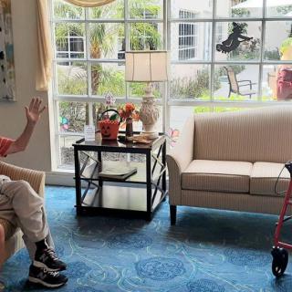 An elderly person sitting in a living room with a walker nearby and sunlight streaming through the window.