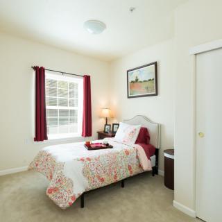 A cozy bedroom featuring a single bed with patterned bedding, a bedside lamp, and a window with red curtains.