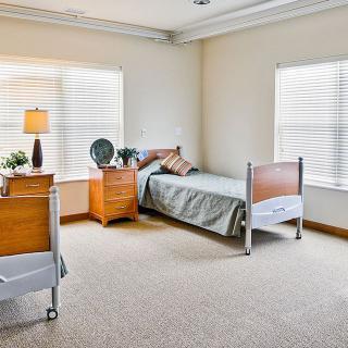 A clean and well-lit bedroom with a single hospital bed, wooden furniture, and large windows.