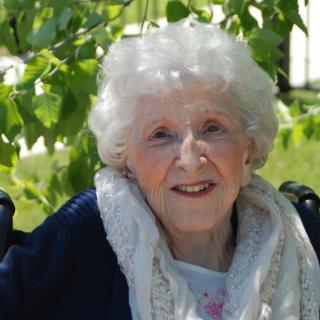 An elderly woman smiling outdoors in a sunny, leafy setting.
