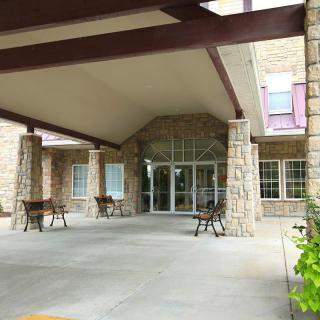 The image shows the entrance to a building with a covered walkway supported by stone pillars, accompanied by outdoor chairs.