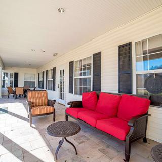 A covered patio with red and orange outdoor furniture, tables, and windows with shutters.