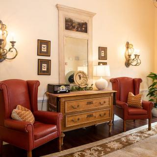 A cozy living room features vintage red armchairs and a wooden dresser with decorative items, enhanced by elegant wall sconces and a large mirror.