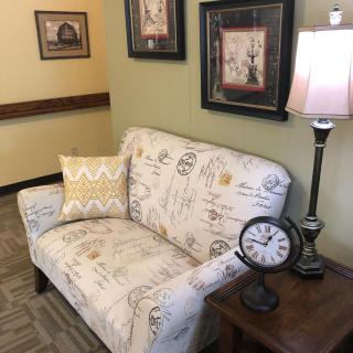 A cozy sitting area featuring a patterned sofa, decorative pillow, table lamp, and a vintage clock, complemented by wall art.