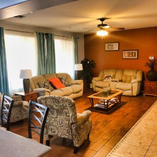 A cozy living room featuring comfortable sofas, armchairs, and a wooden coffee table. The space is warmly lit and complemented by long curtains and a ceiling fan.