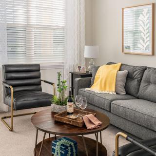 A cozy living room featuring a gray sofa with a yellow throw, a black armchair, and a round wooden coffee table adorned with decor items. The space is completed with wall art, a lamp, and green plants.