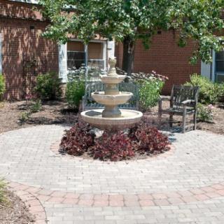 A serene garden scene featuring a central fountain surrounded by paved walkways and chairs, with lush greenery and a tree providing shade.