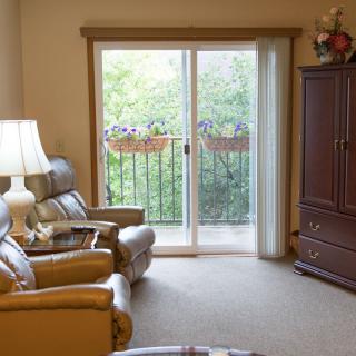 A cozy living room featuring armchairs, a lamp, and a sliding glass door leading to a small balcony with plants.