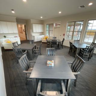 A modern communal dining area featuring multiple tables and chairs, a kitchenette, and large windows.