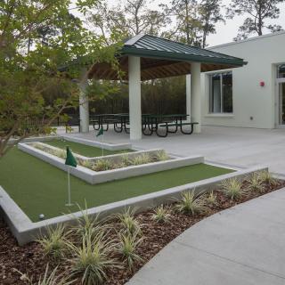 An outdoor mini-golf area with artificial turf, sheltered seating, and surrounding greenery.