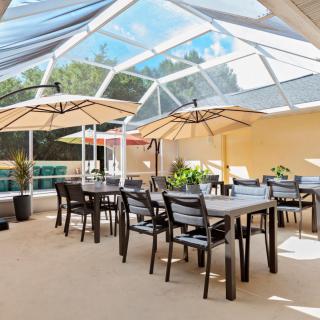 A bright outdoor patio dining area with tables and chairs under large umbrellas, covered by a glass roof.
