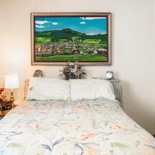 A cozy bedroom featuring a neatly made bed with floral bedding, flanked by lamps. A framed painting of a landscape hangs on the wall above the headboard.