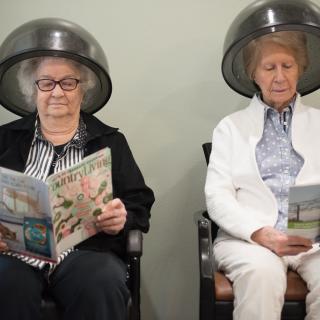 Two elderly women sitting under hair dryers in a salon, reading magazines.