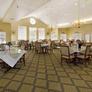 A spacious dining area with elegantly arranged tables and chairs, illuminated by chandeliers and natural light from large windows.