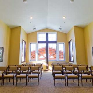 A serene chapel interior with rows of chairs and a large cross in front of tall windows.