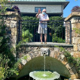 A person stands on a stone bridge above a fountain in a garden setting.